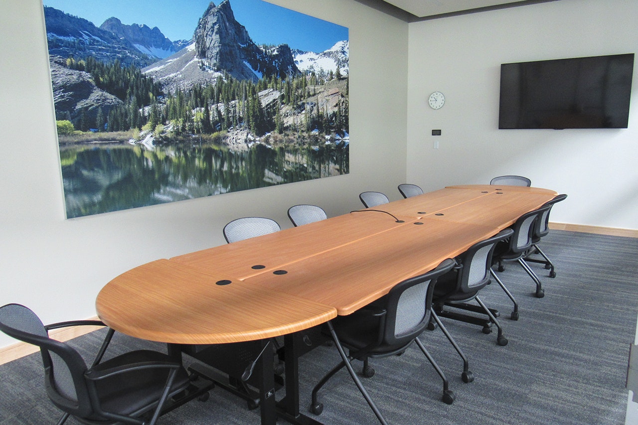 A modern conference room with a long table, chairs, a scenic wall canvas, a clock, and a flat-screen TV.