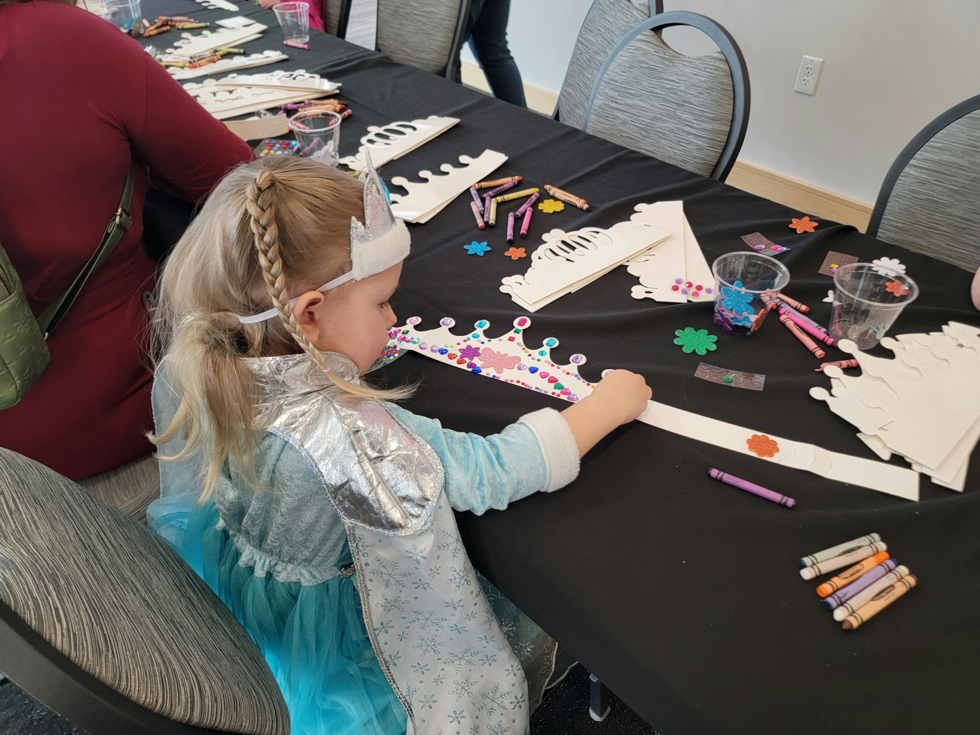 A child in a costume decorates a crown with stickers at a craft table with crayons and paper crowns.