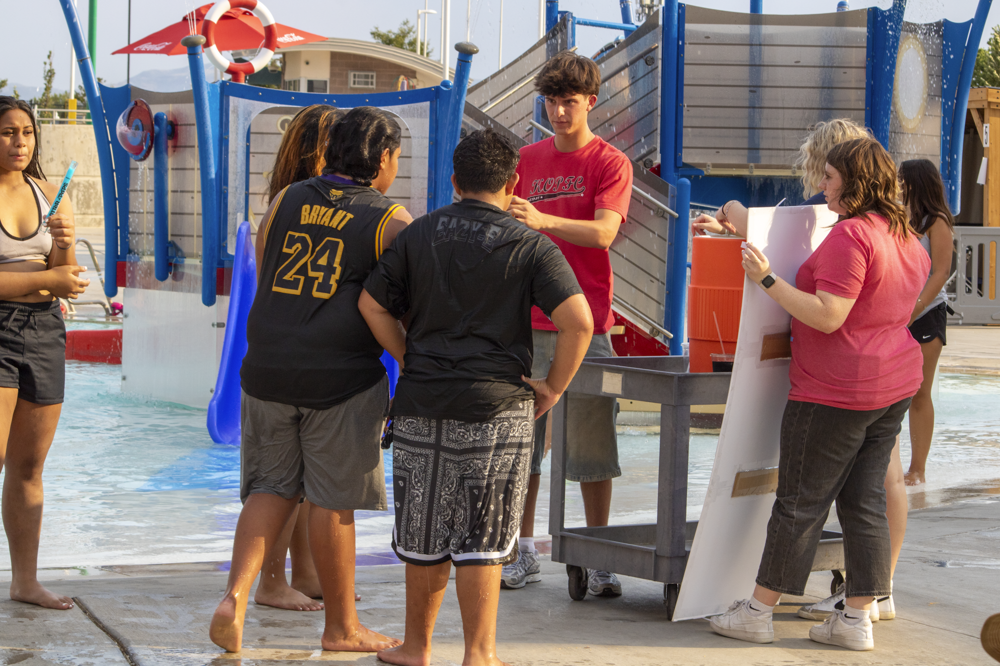 A group of people is gathered by a splash pad, engaged in conversation, with some holding signs and wearing casual summer attire.