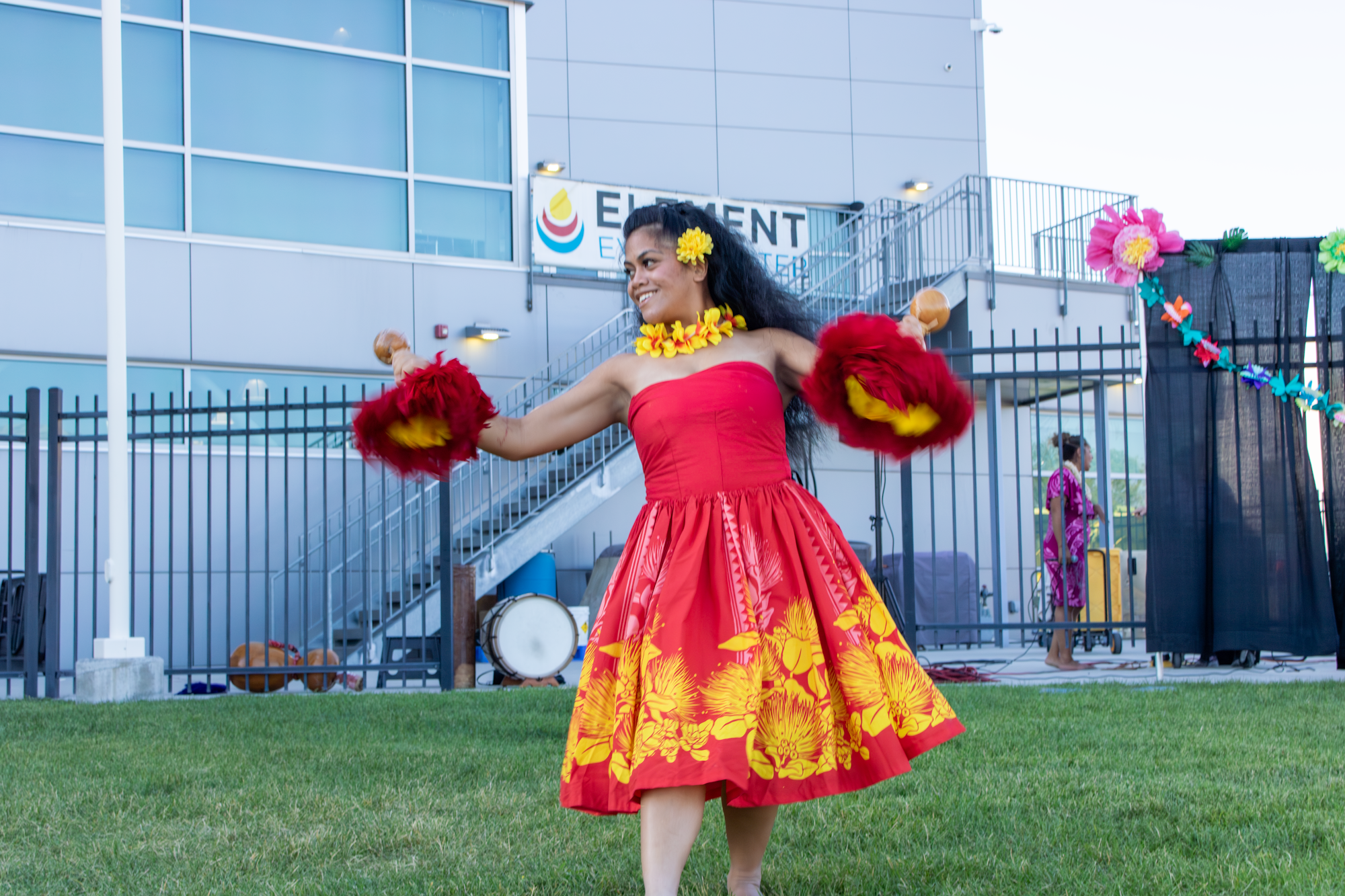 A dancer in a vibrant red dress with floral patterns performs outdoors, holding maracas while surrounded by decorative elements.