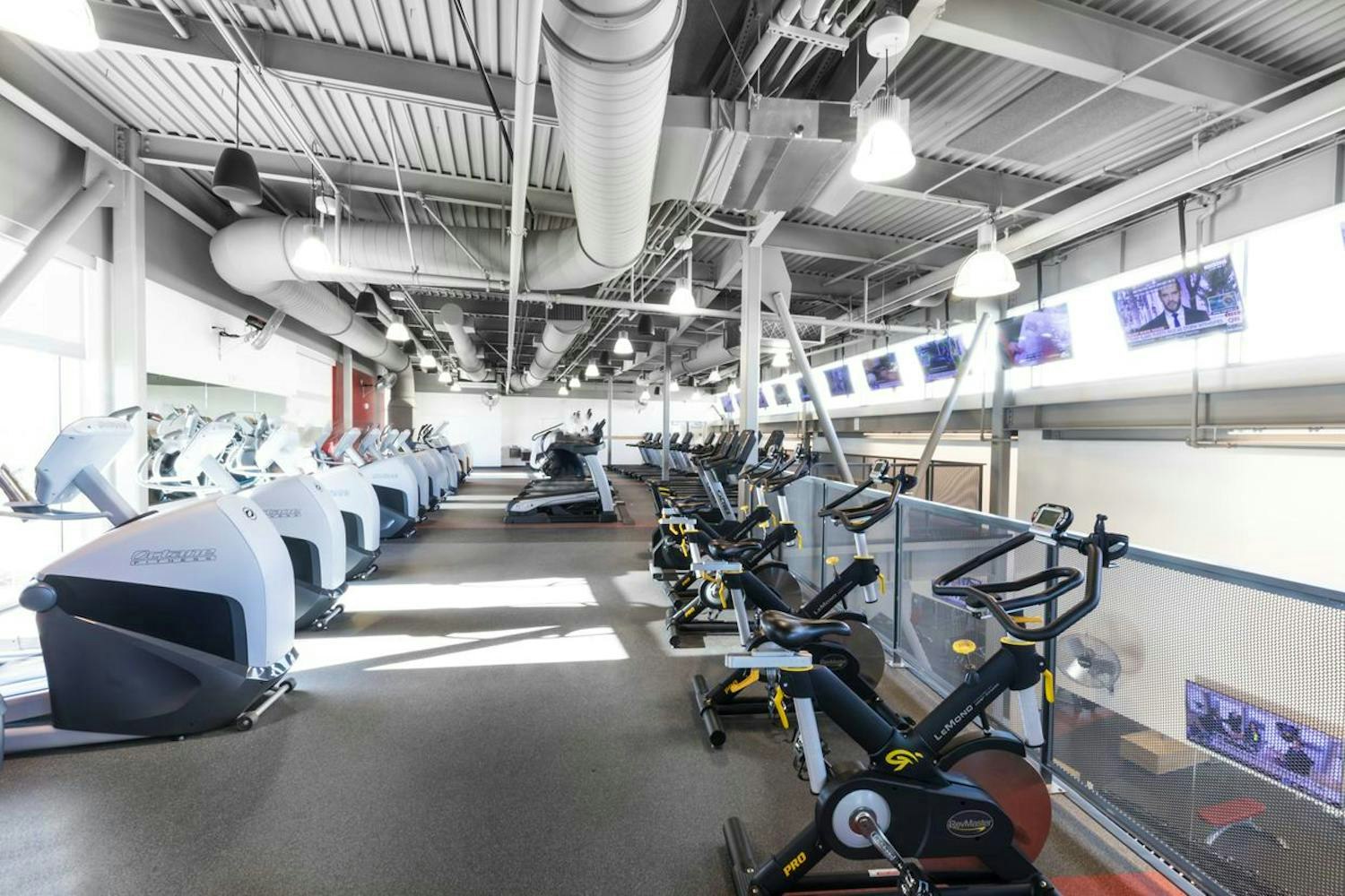 An indoor gym with rows of exercise bikes and treadmills, bright lighting, TVs, and industrial-style ceilings.