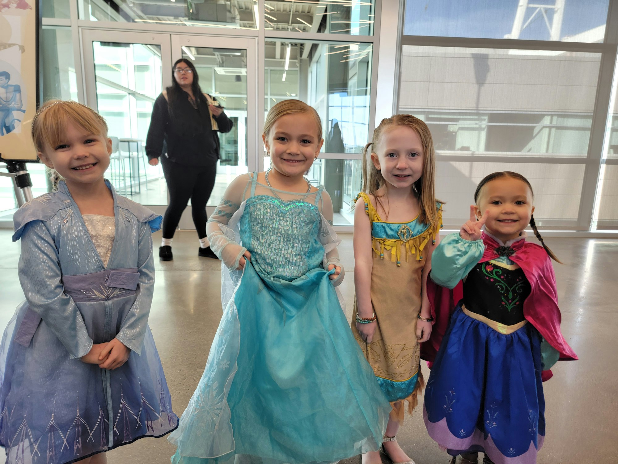 Four children dressed as princesses posing indoors, with an adult in the background.
