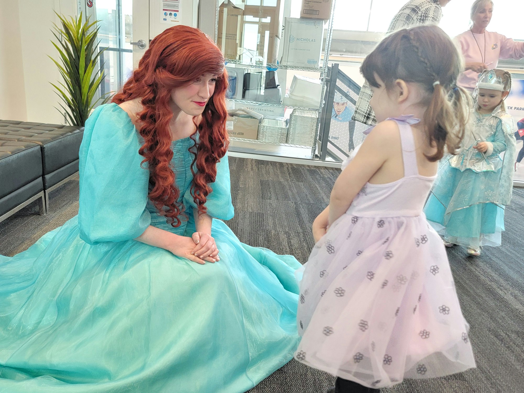 A character in a turquoise dress interacts with a young girl in a lavender dress, surrounded by other kids in costumes.