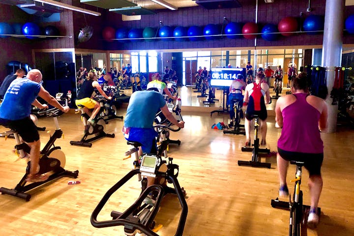 Indoor cycling class with participants on stationary bikes, exercise equipment, and a timer displaying 00:18:14.