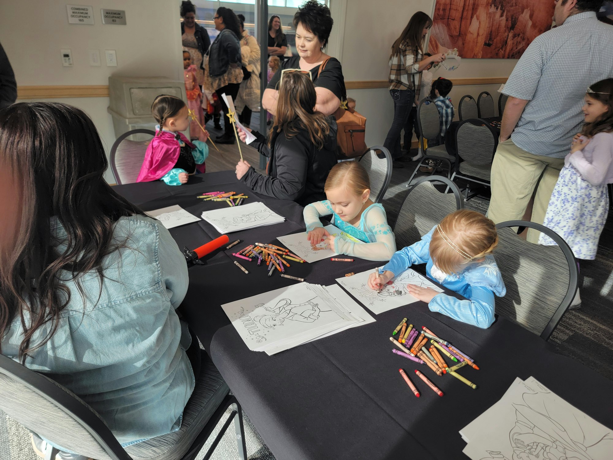 Children in costumes coloring pictures at a table with crayons, surrounded by adults and other kids.