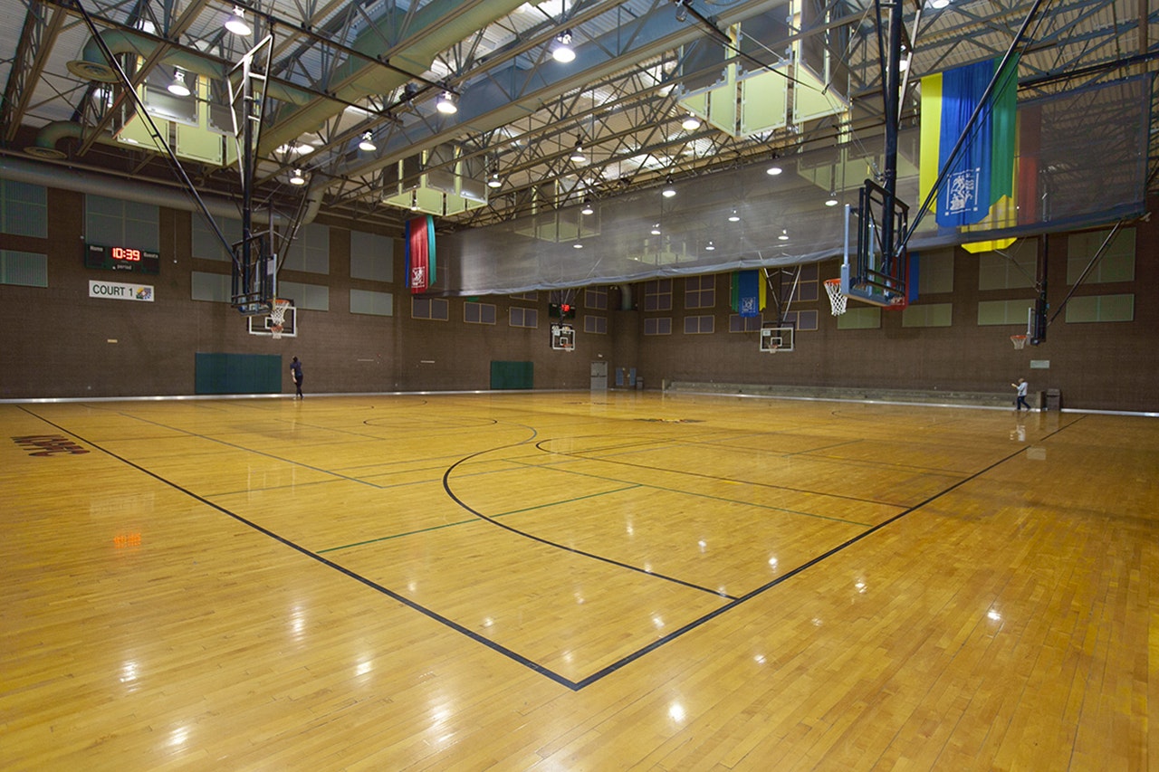 An indoor basketball court with a polished wooden floor, hoops at each end, and banners on the wall.