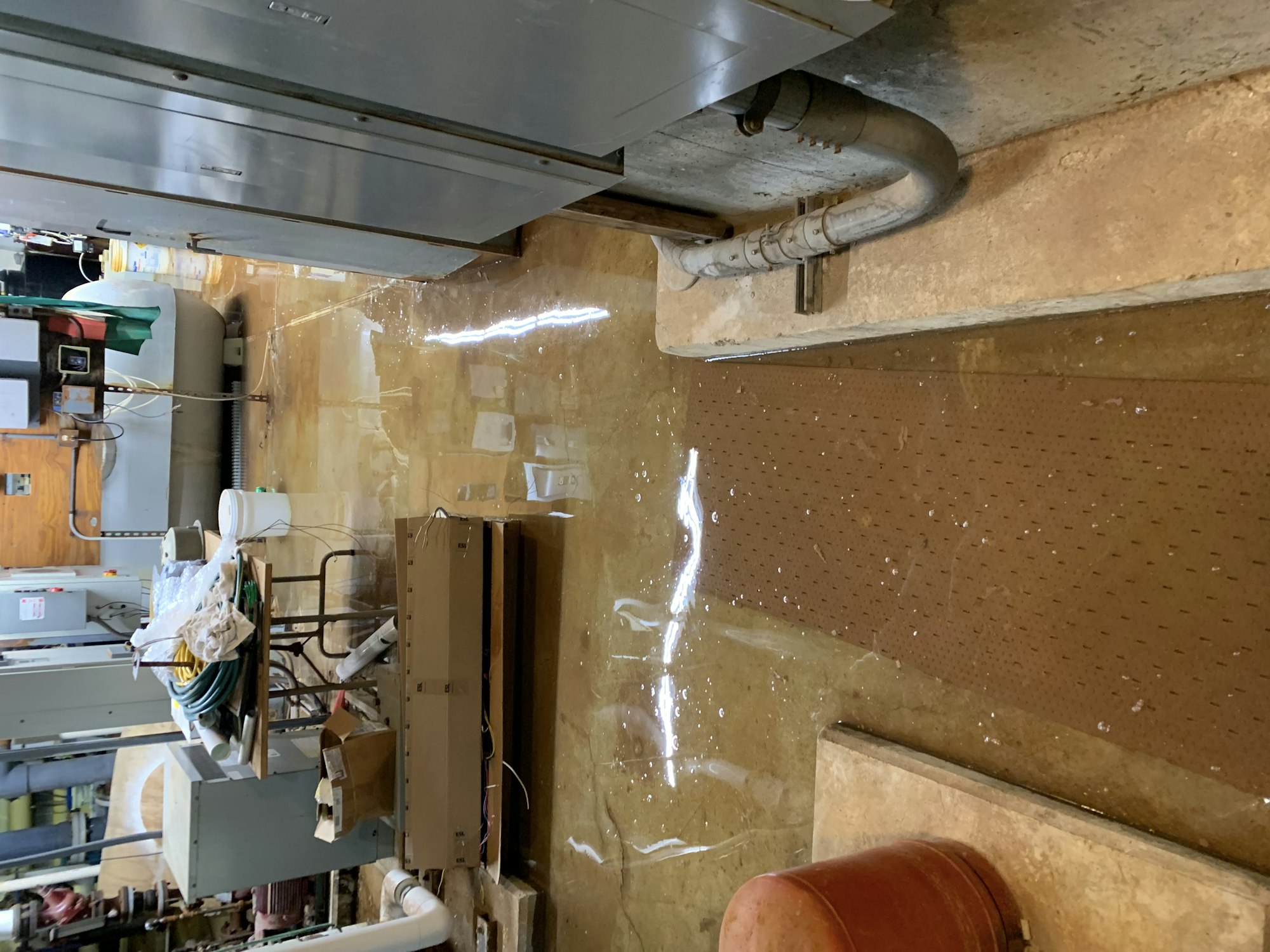A flooded industrial space with standing water on the floor, electrical panels, pipes, and various tools scattered around.