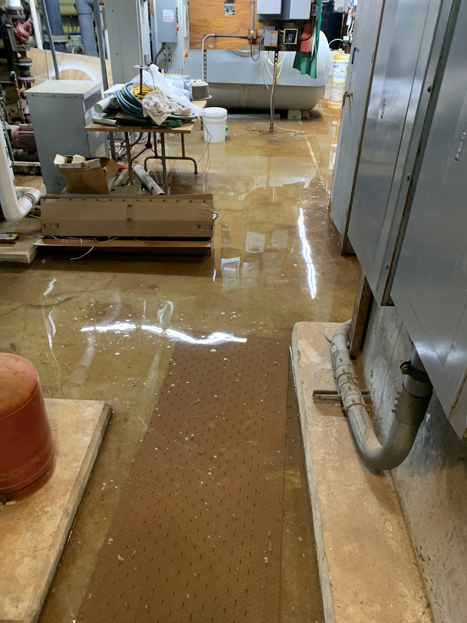 A flooded industrial space with standing water on the floor, electrical panels, pipes, and various tools scattered around.