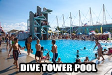 A busy outdoor pool with a diving tower, people swimming and lounging under the sun.