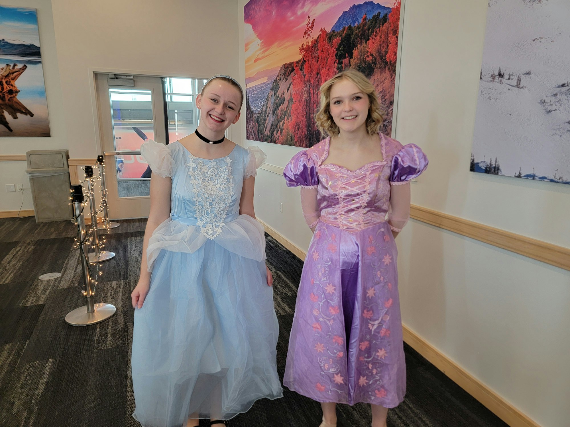 Two people in princess costumes, one in blue and one in purple, standing in a room with scenic wall art.