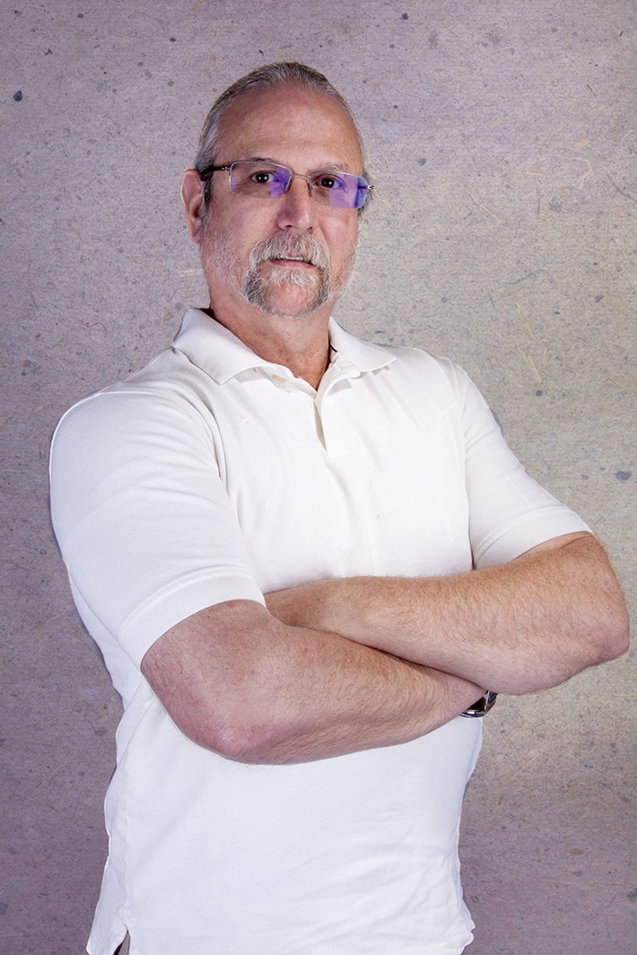 A person with glasses and a mustache, wearing a white polo shirt, standing with arms crossed.