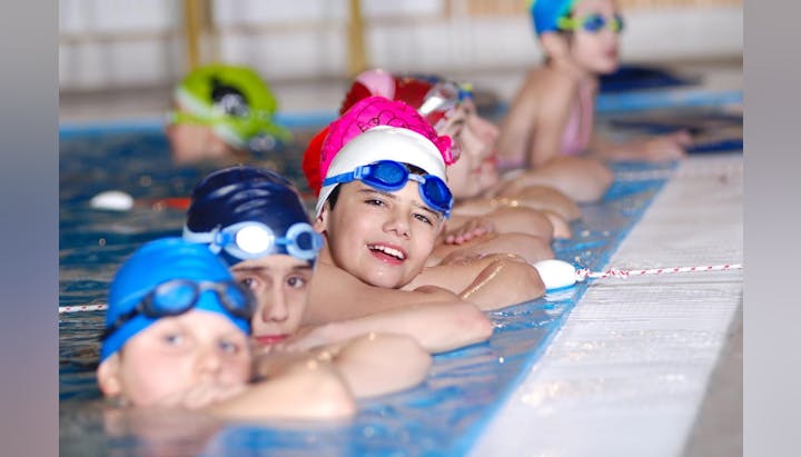 Children in swim caps and goggles at a pool edge, ready to swim.