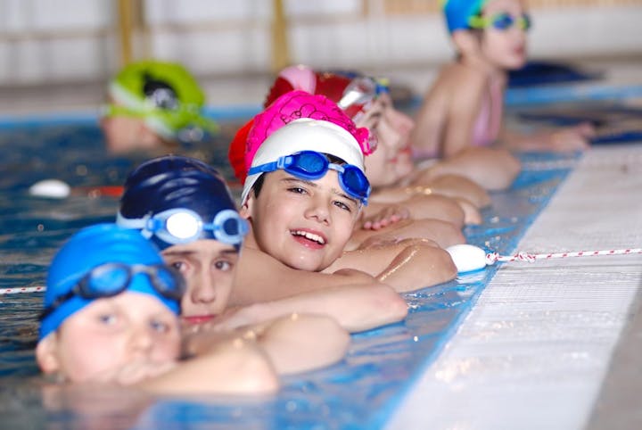 Children in swim caps and goggles at a pool edge, ready to swim.