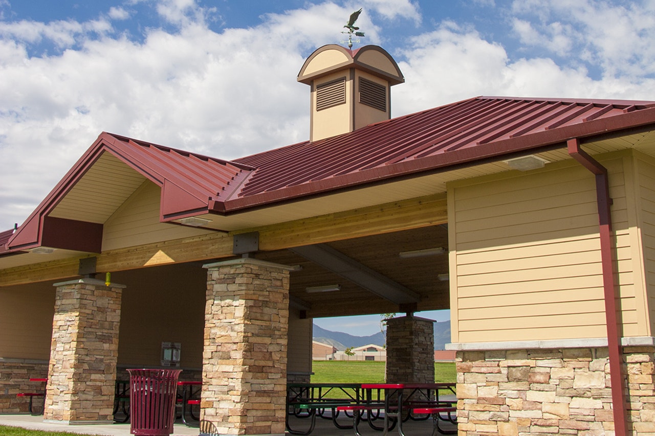 Pavilion with red metal roof, stone pillars, picnic tables, weathervane atop, set against a blue sky with clouds.