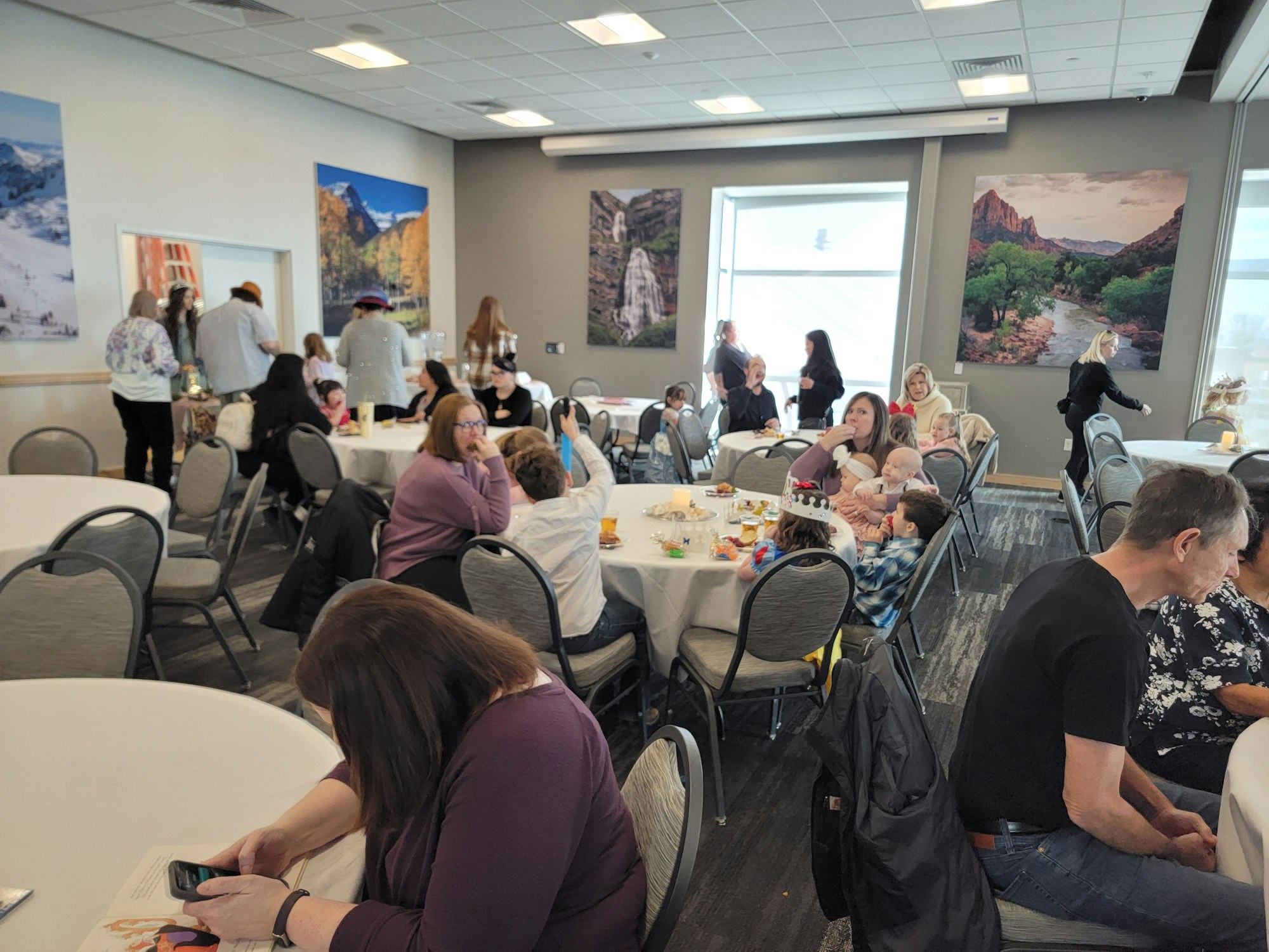 A gathering in a room with round tables and people socializing, some using phones. Large scenic photos are on the walls.