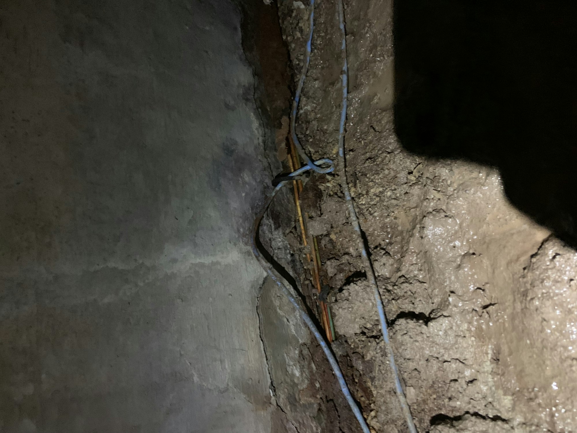 A rough wall with exposed electrical wiring and cables tied together, possibly in a basement or underground area.