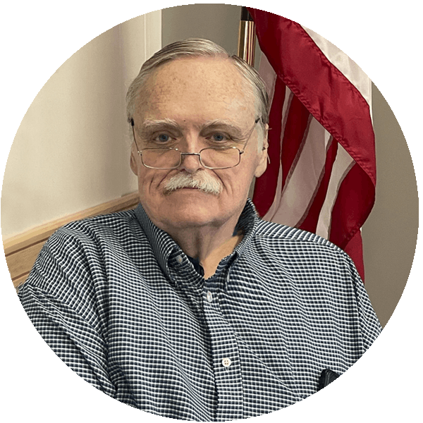 A man with a mustache wearing glasses and a checkered shirt, with an American flag in the background.