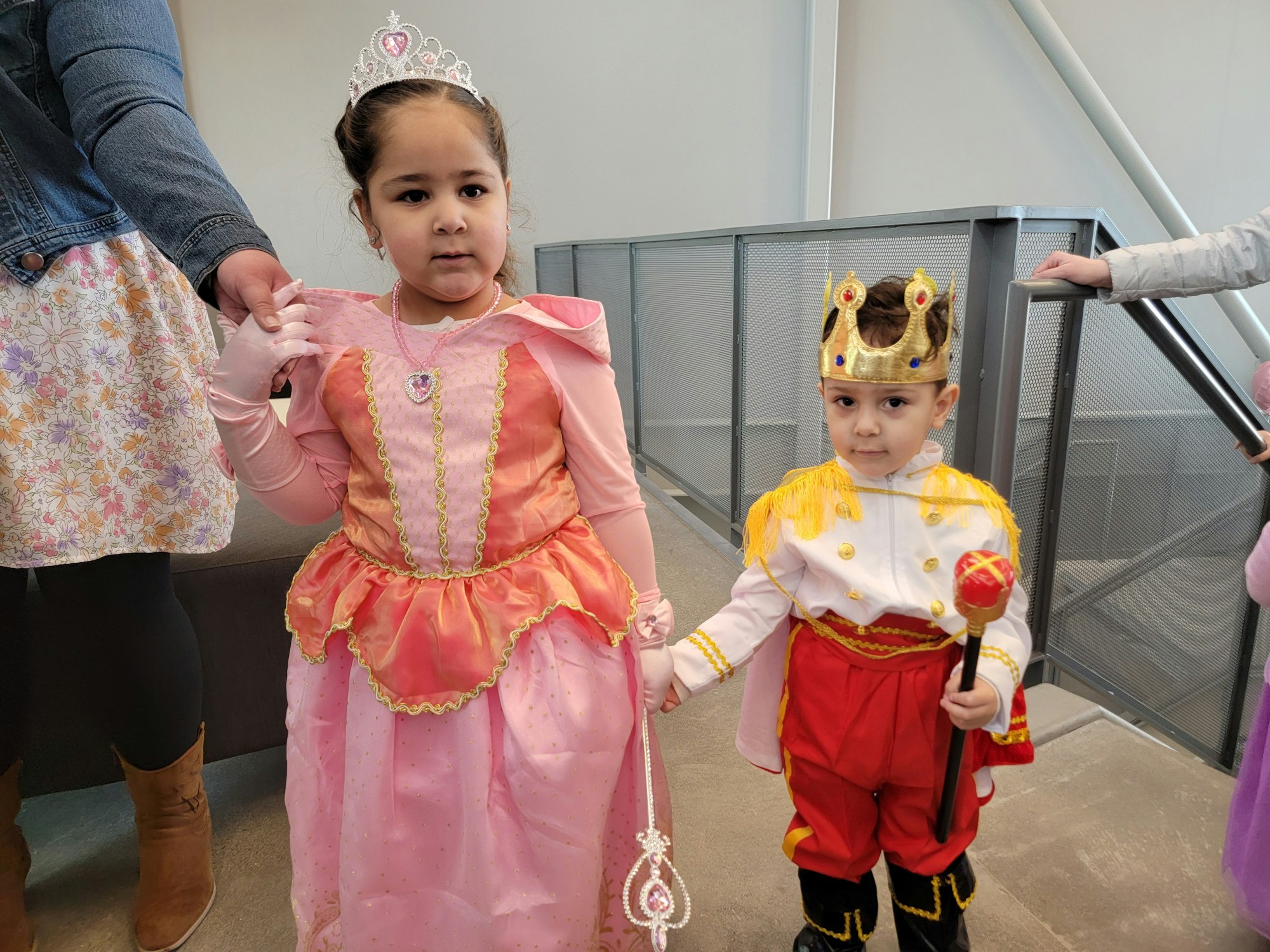 Two children dressed as a princess and a king, holding hands and being escorted by adults.