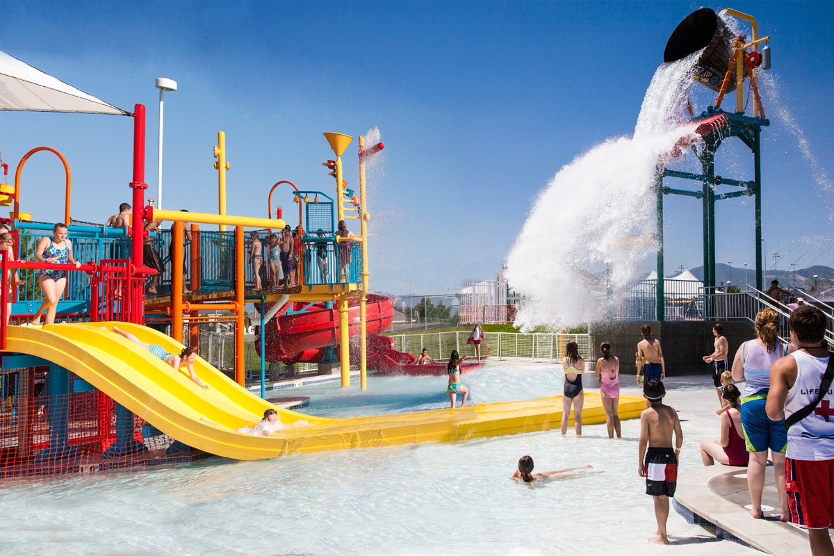 A vibrant waterpark scene with kids sliding and playing, a large splash from a water bucket, and onlookers enjoying a sunny day.