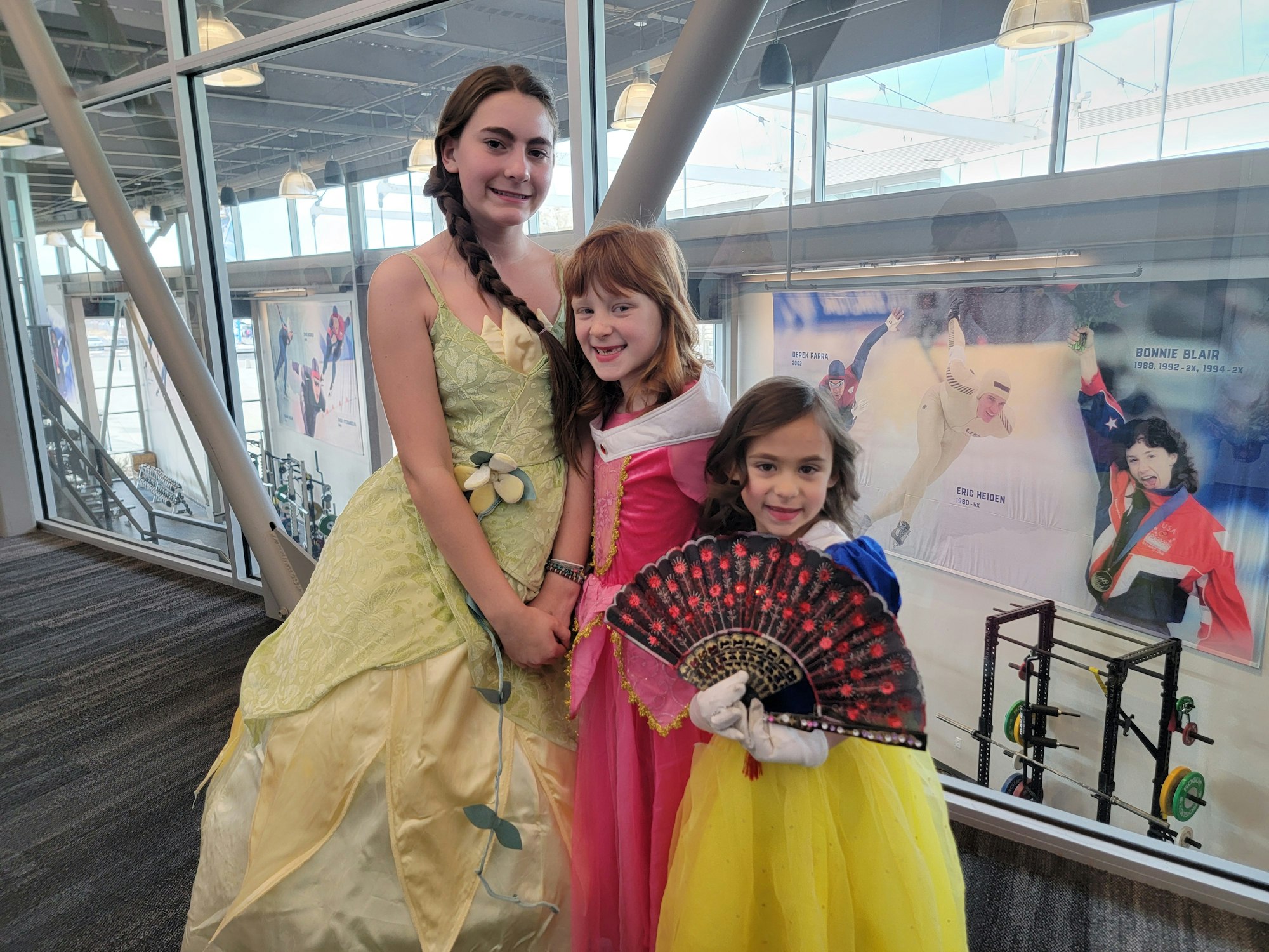 Three children in princess costumes posing in a building with photo displays and weightlifting equipment.
