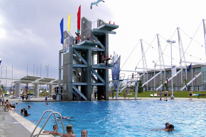 Outdoor pool with diving platforms, swimmers, flags, and a modern building in the background.