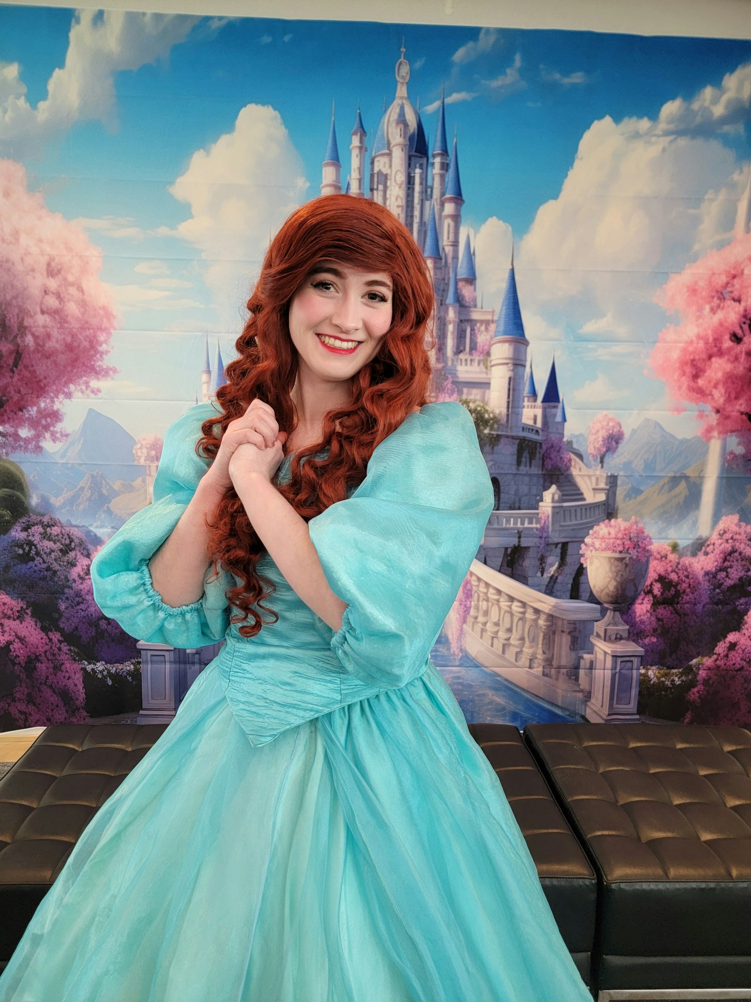 A person in a turquoise dress with red hair, posing in front of a fantasy castle backdrop.