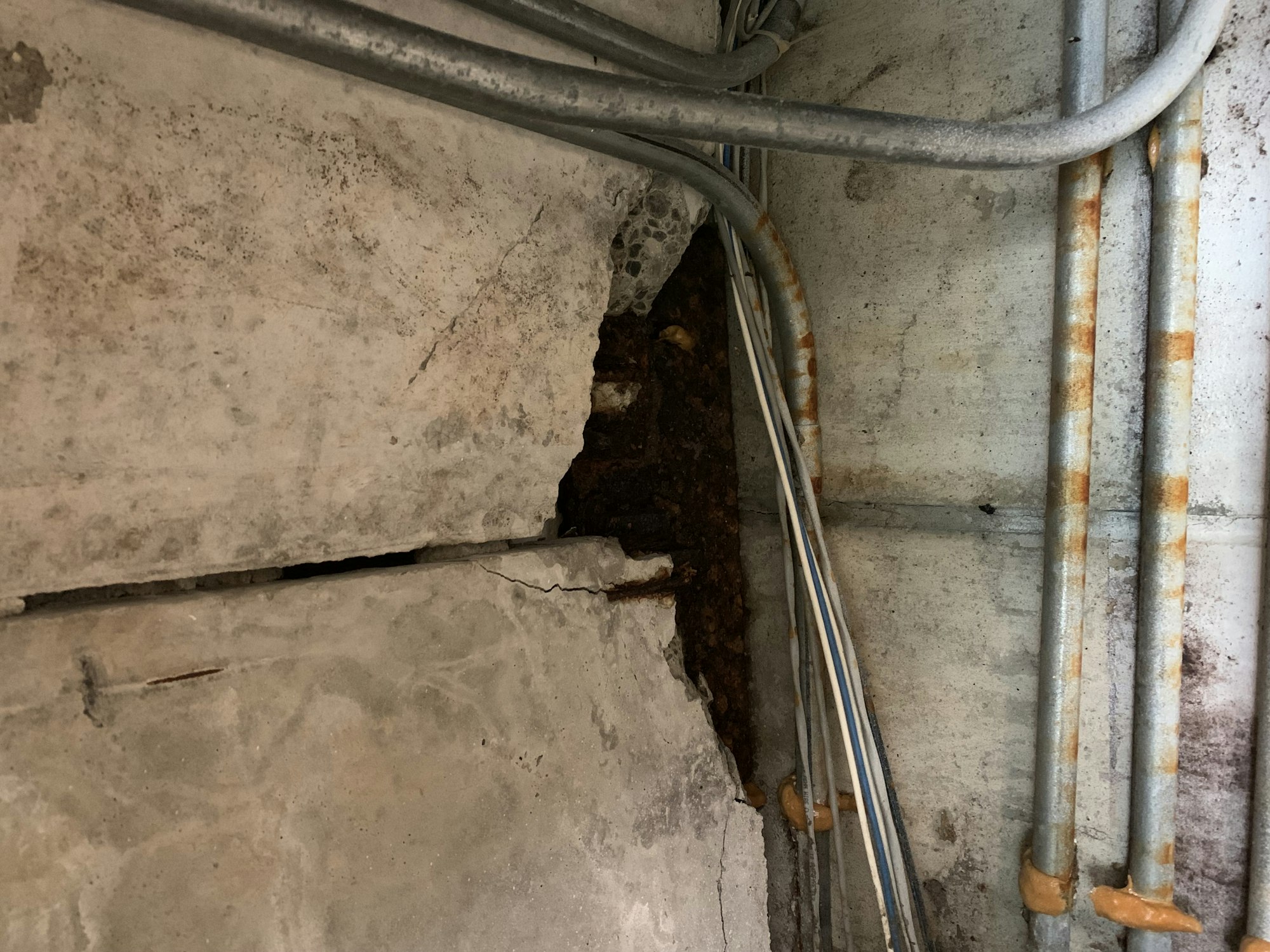 Cracked concrete wall with exposed cables and rusted metal pipes.