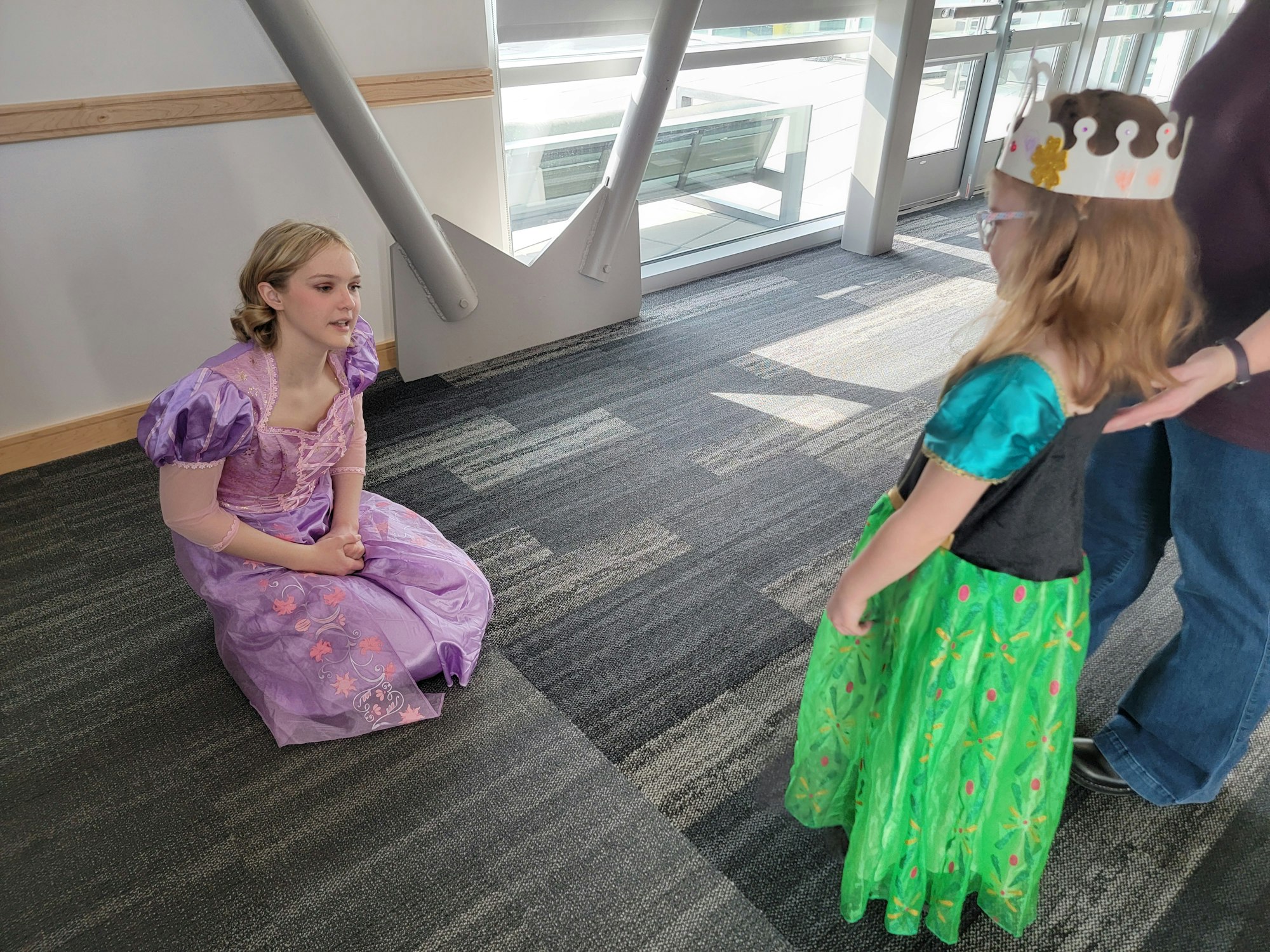 A person in a purple dress kneels, facing a child in a green dress and crown.