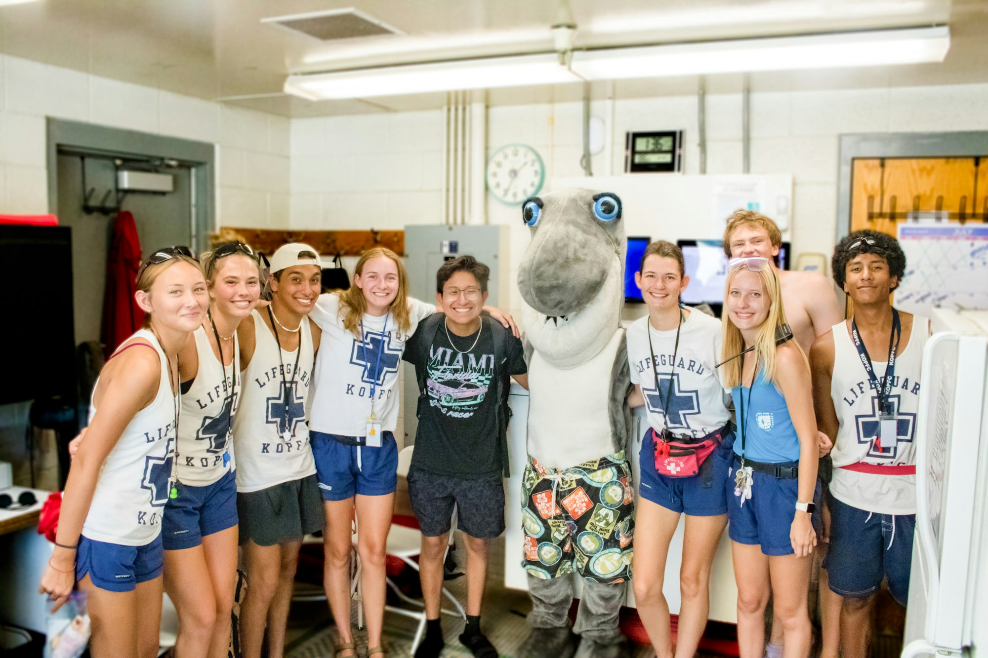 A group of lifeguards posing with someone in a shark costume indoors.