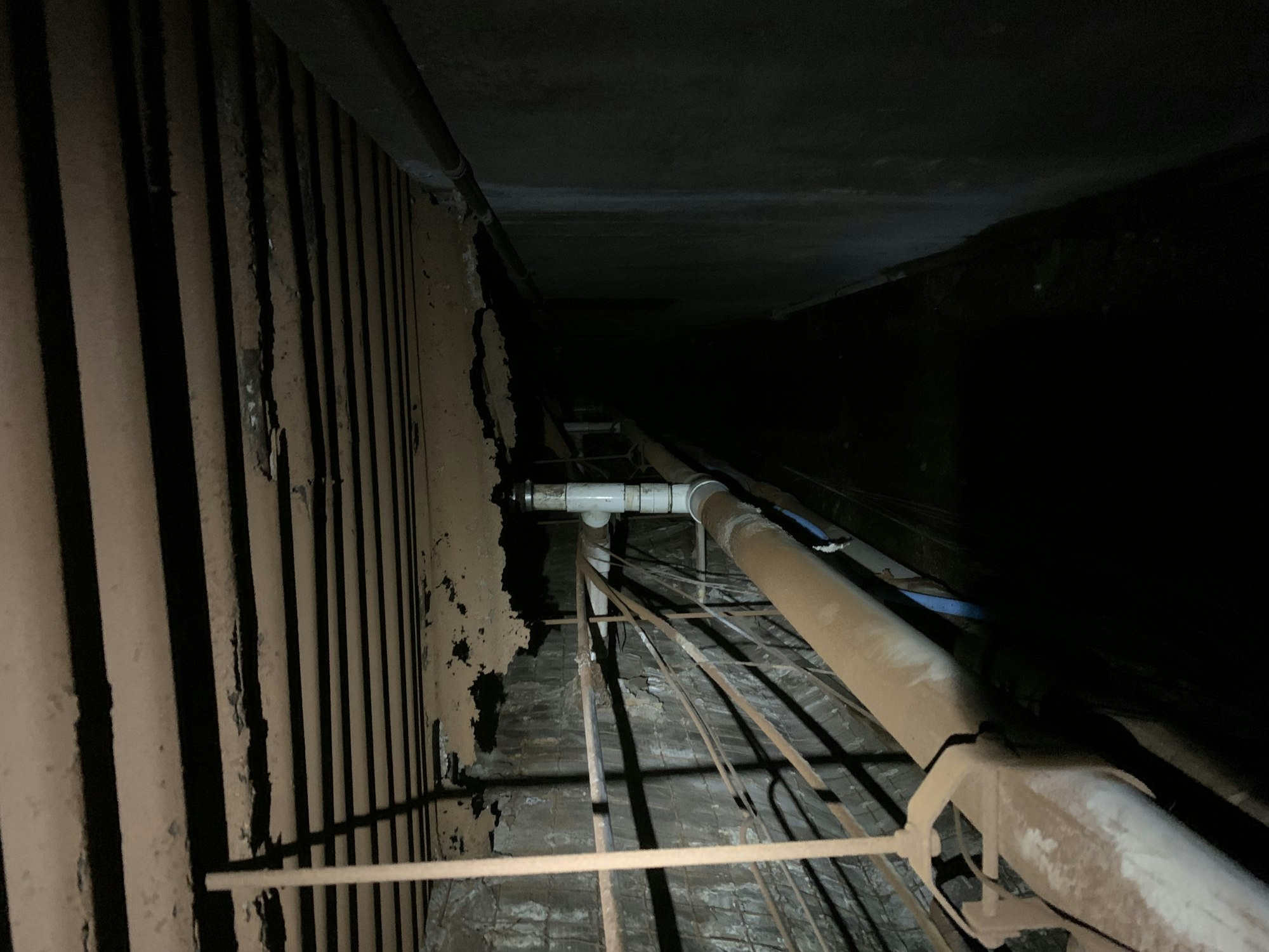 Dark, narrow space with rusty pipes, wires, and peeling walls.