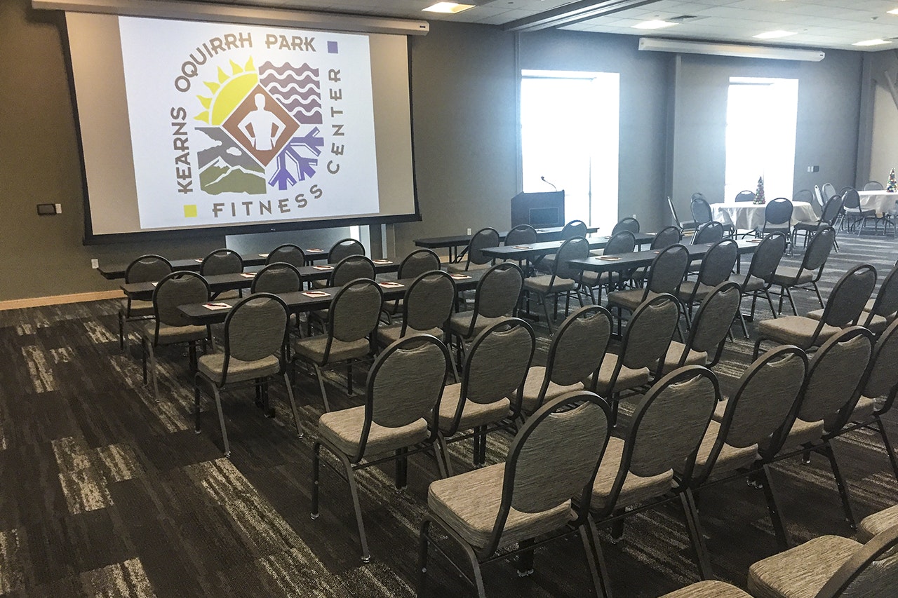 Conference room with rows of chairs facing a projection screen displaying a logo, podium on side, and tables with decor in background.