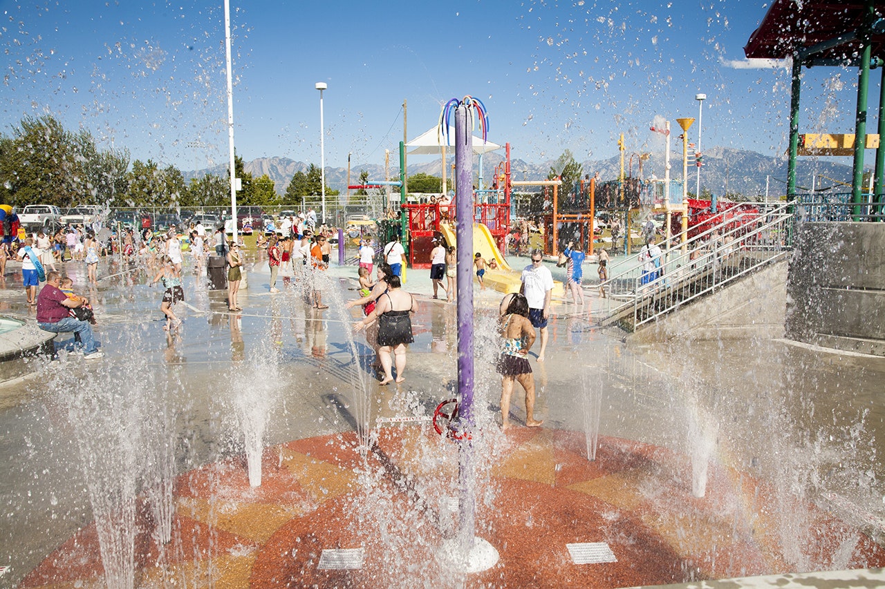 A splash pad with children playing, water jets, and a playground in the background, on a sunny day.