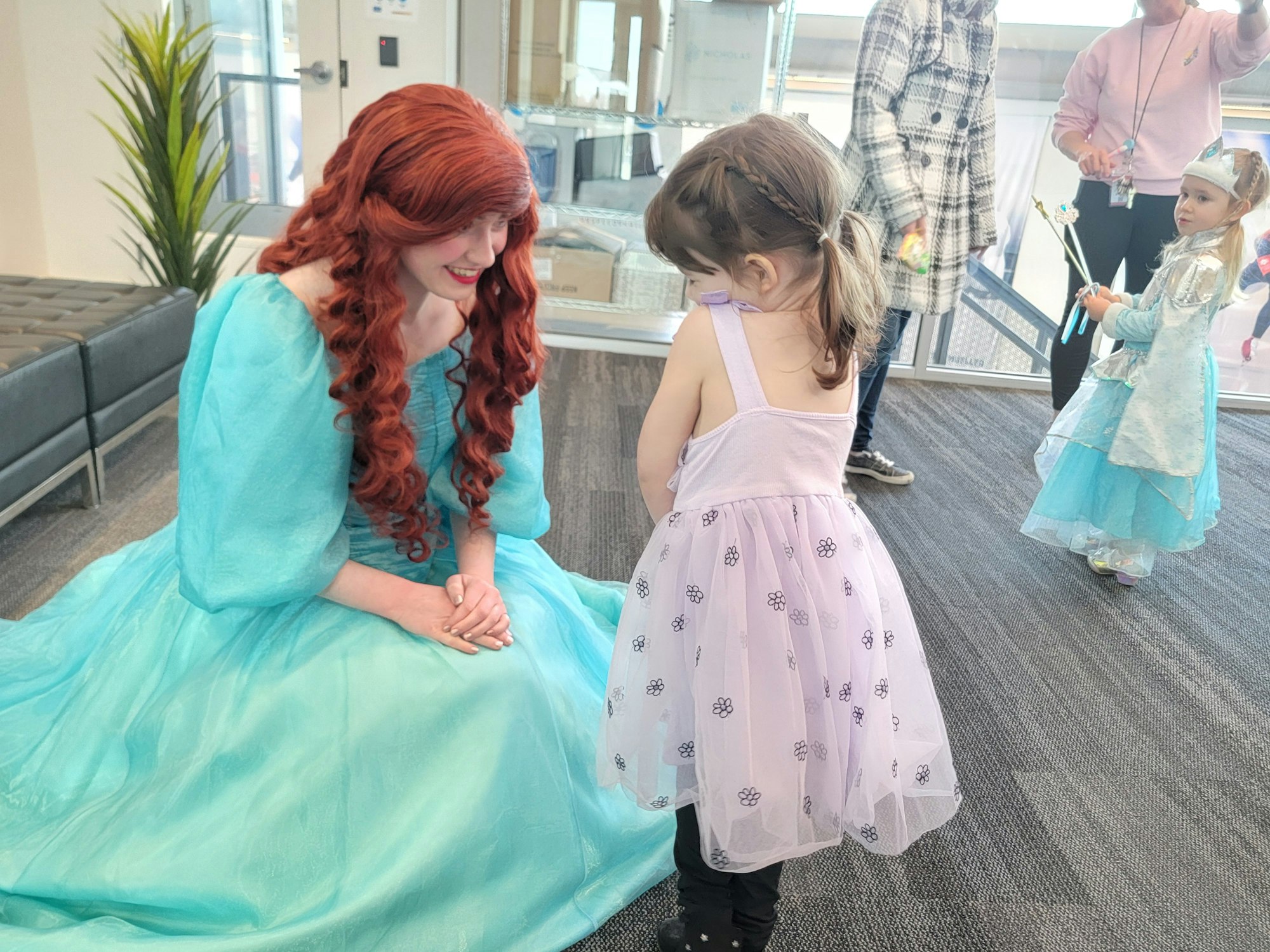 A woman in a turquoise dress interacts with a child in a lilac dress. Another child dressed as a fairy is nearby with adults.