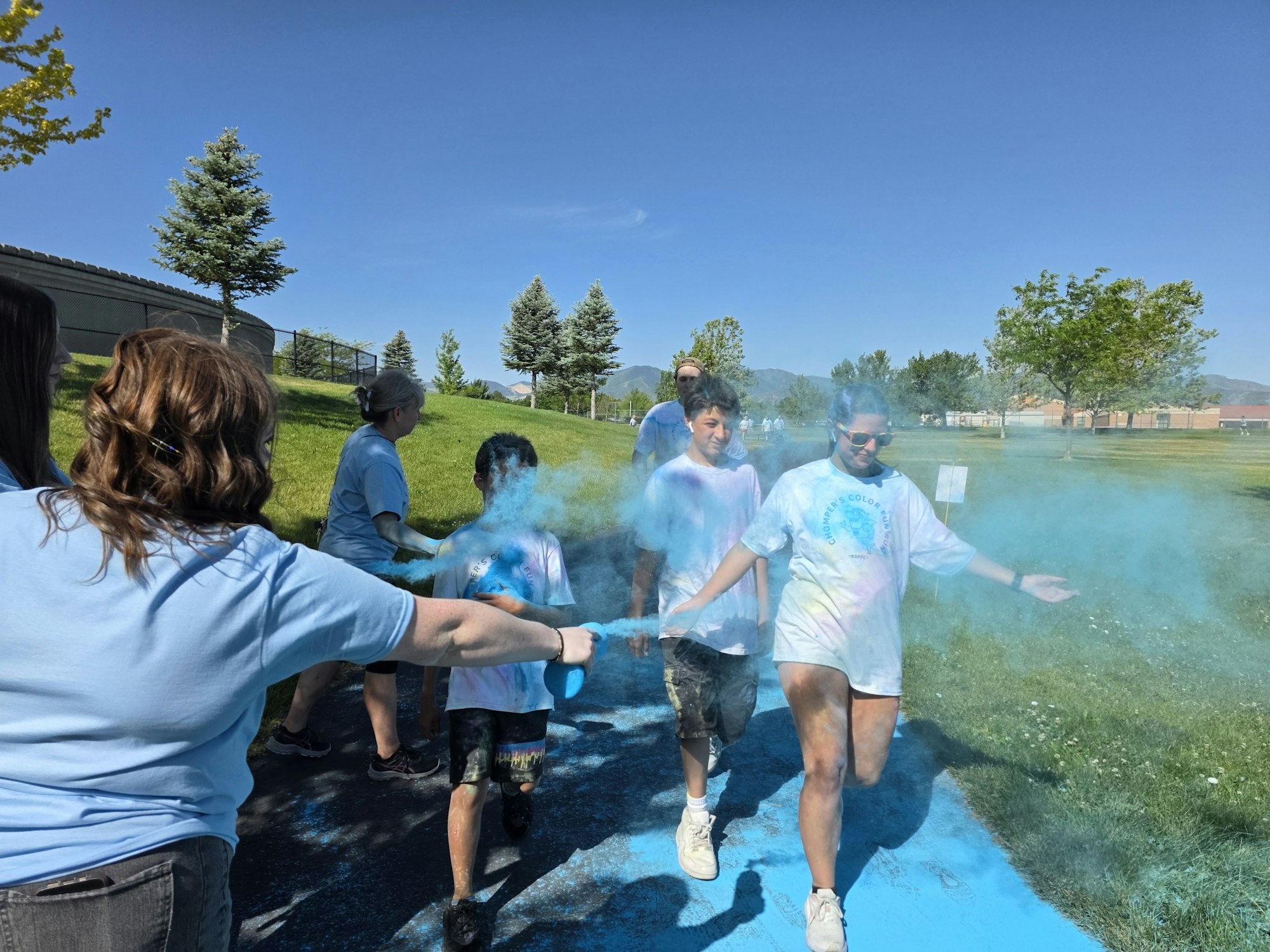 A group of kids and adults participate in a color run, throwing vibrant colored powder in a sunny outdoor setting.