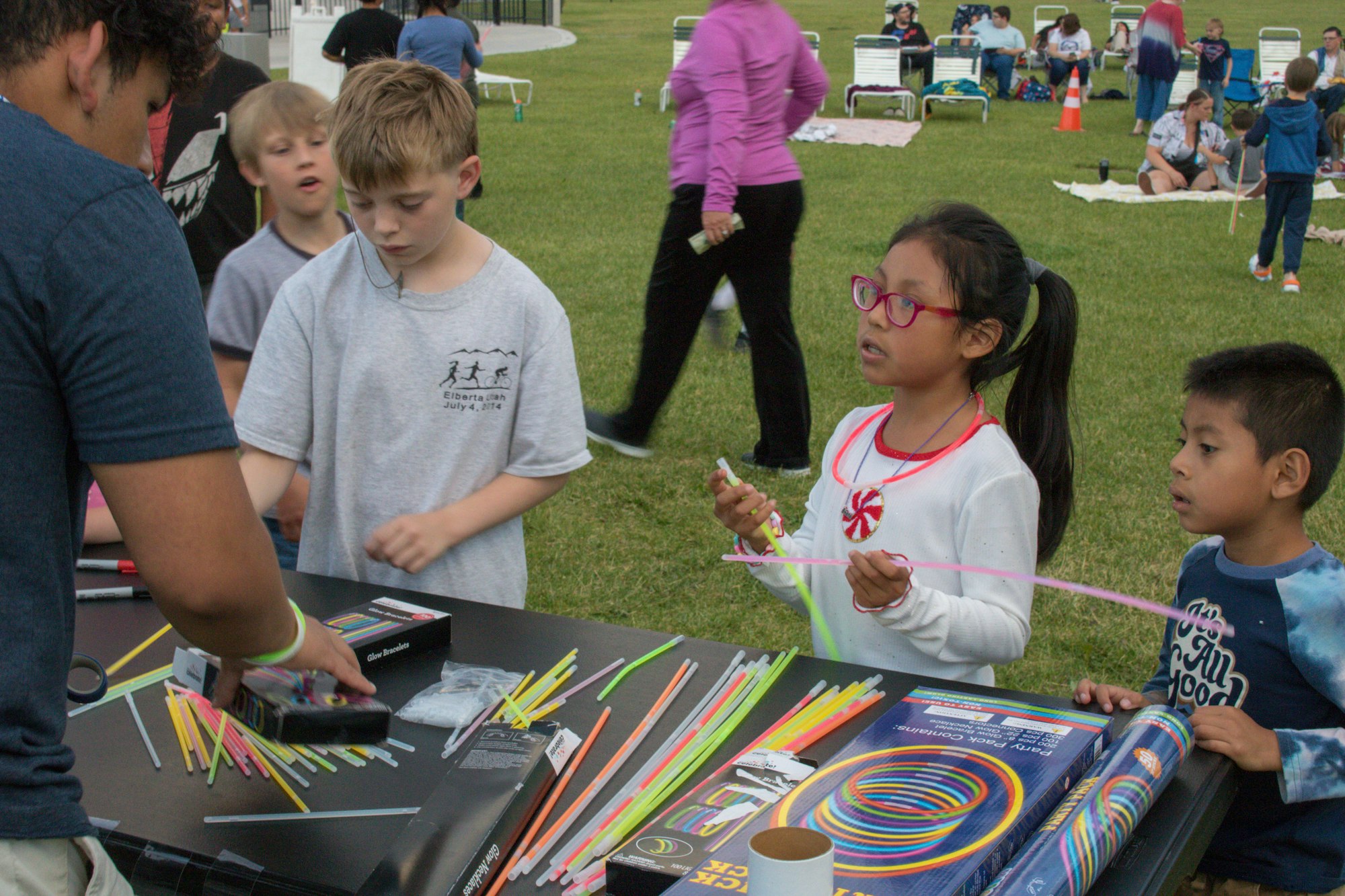 Children at an outdoor event with glow sticks and a man assisting. People in the background are on a lawn.