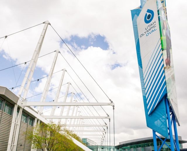 Modern building with a white structural frame and a blue banner reading "Utah Olympic Oval" under a cloudy sky.