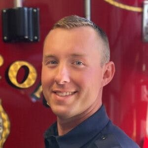 A person in uniform is smiling, with a red background that appears to be firetruck-related equipment.