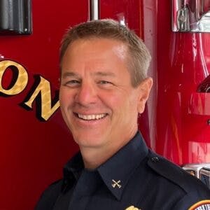 A person in a black uniform is smiling against a red background.