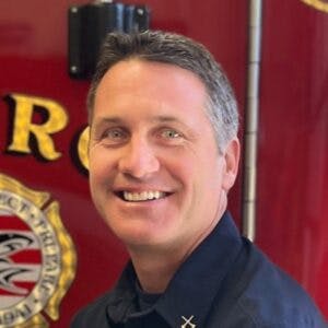 A smiling man in uniform stands before a fire department logo on a red background.