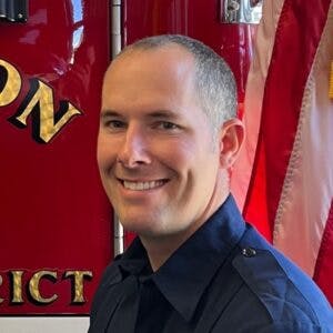 A person in a uniform smiling in front of a fire truck and an American flag.