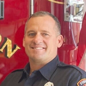 A person in a dark uniform stands smiling in front of a red fire truck.