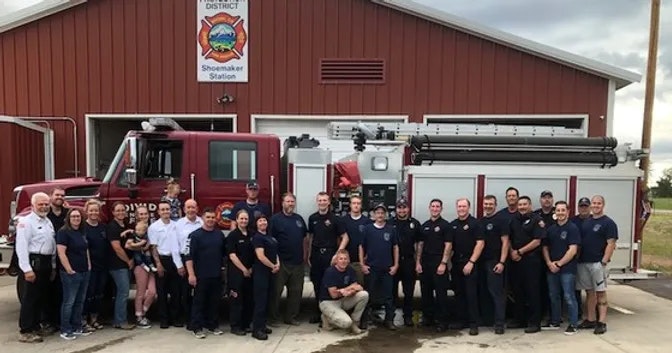 A group of firefighters posing in front of a fire station and trucks.