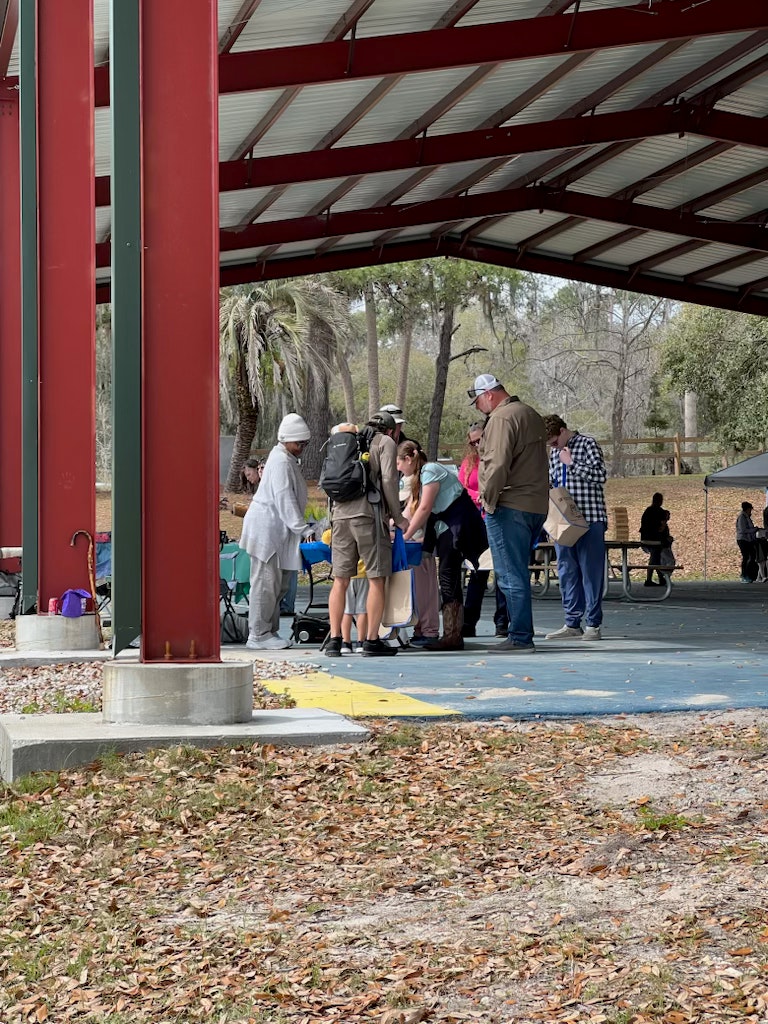 People gathered under a shelter, some sitting, others standing in a line, with trees and leaves around.