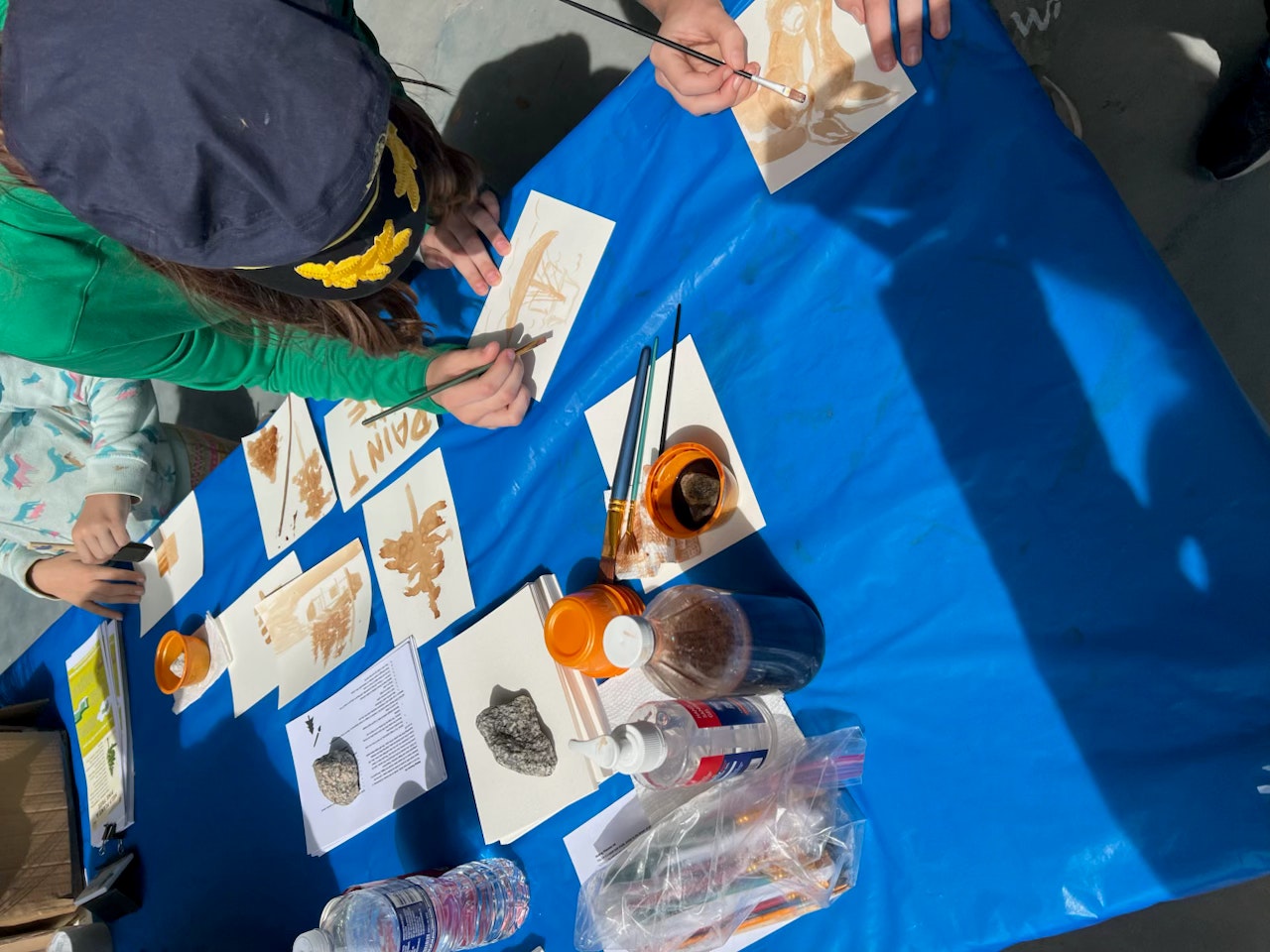 People painting on cards at a table with paint, brushes, and reference images on a blue tablecloth.