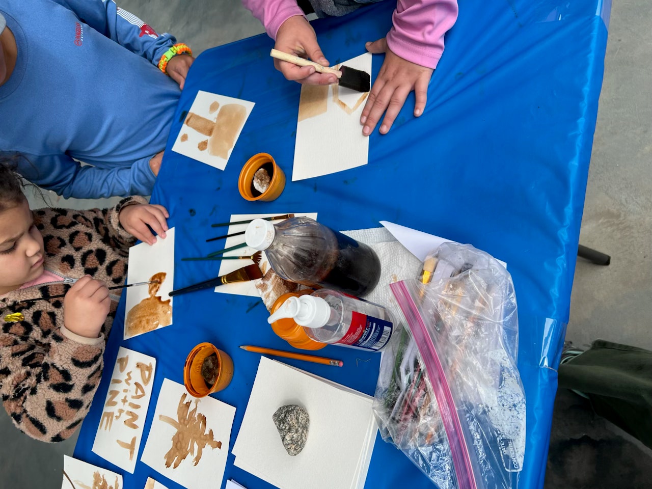 Children painting with natural brown colors at a table covered in a blue cloth, surrounded by art supplies.