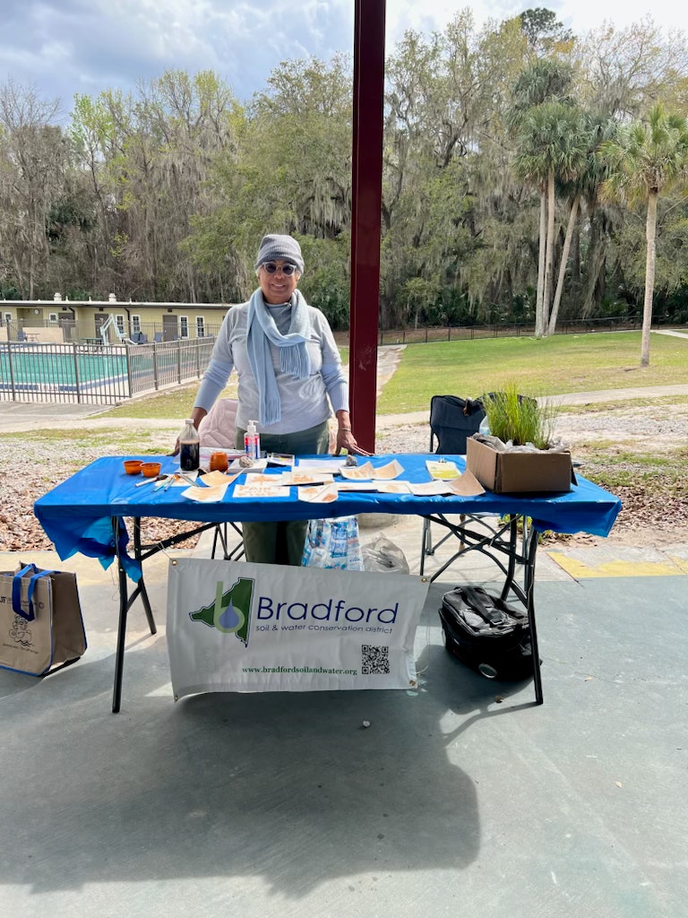 A person stands behind a table with various items under a covered area. There's a "Bradford Soil & Water Conservation District" sign.