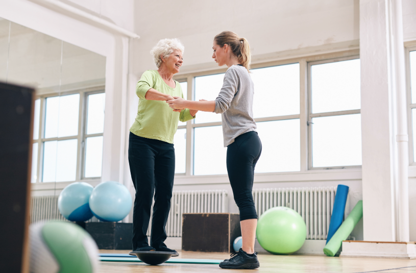 Two women exercising with a balance board in a fitness studio, surrounded by exercise balls and mats.