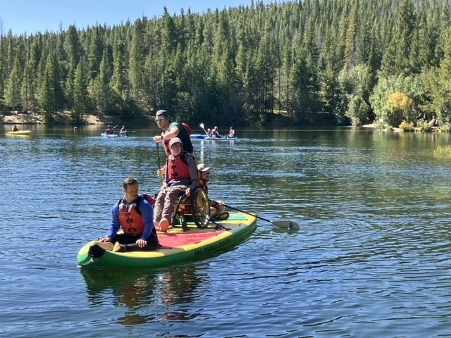 People on a paddleboard in a lake, with trees in the background, enjoying a sunny day.