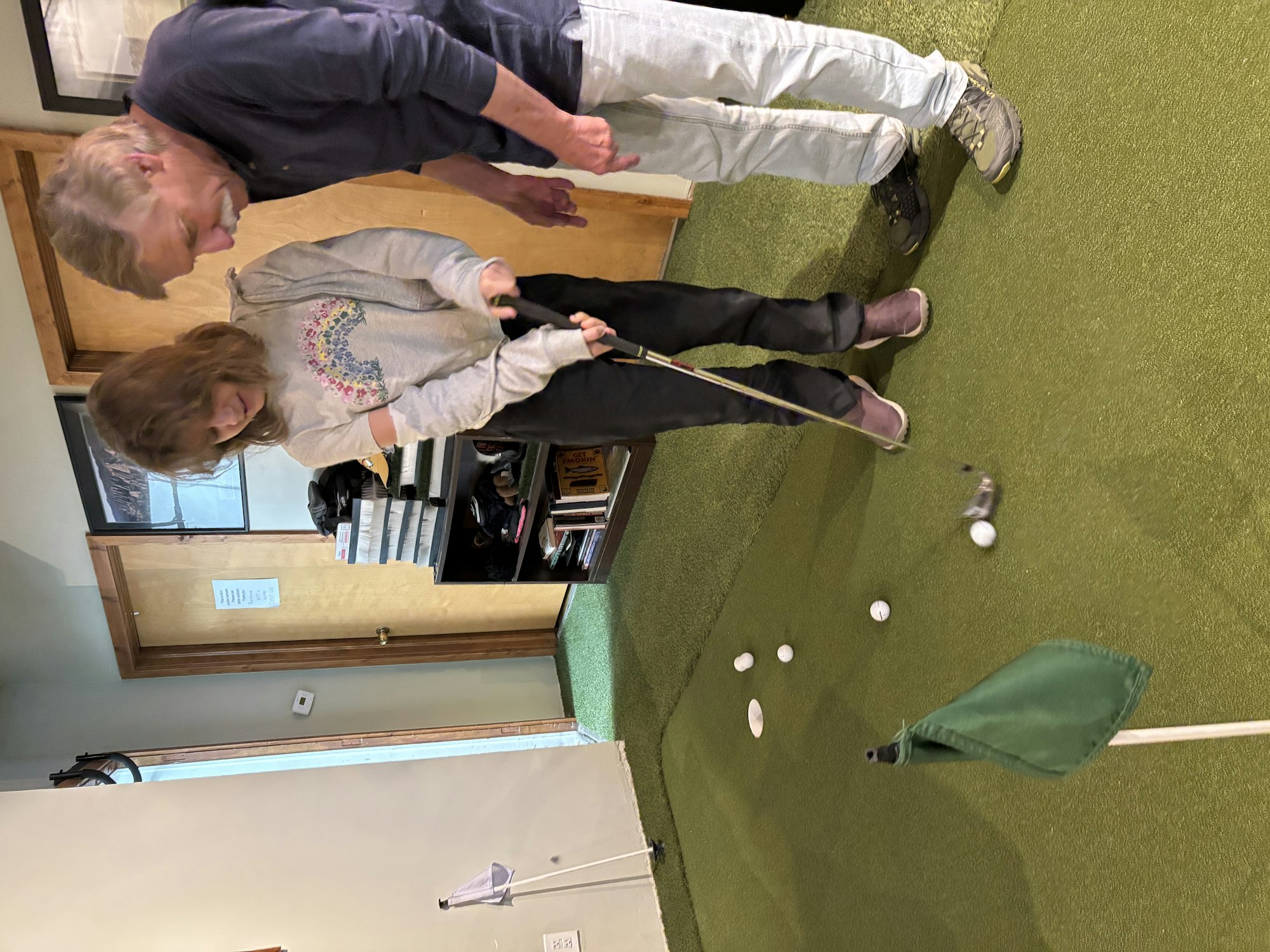 Two people playing mini-golf indoors, focusing on putting near a small green flag.