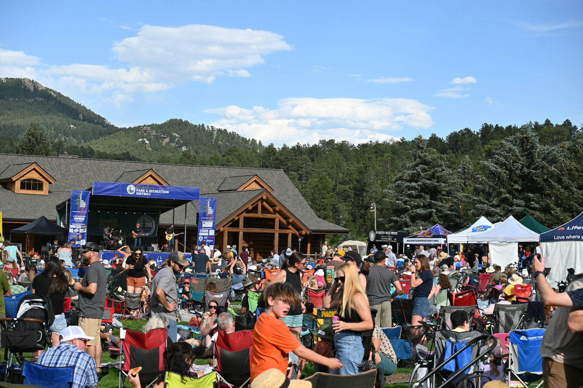 Outdoor concert with a crowd sitting on chairs, tents, and stage with performers, set against a backdrop of trees and mountains.